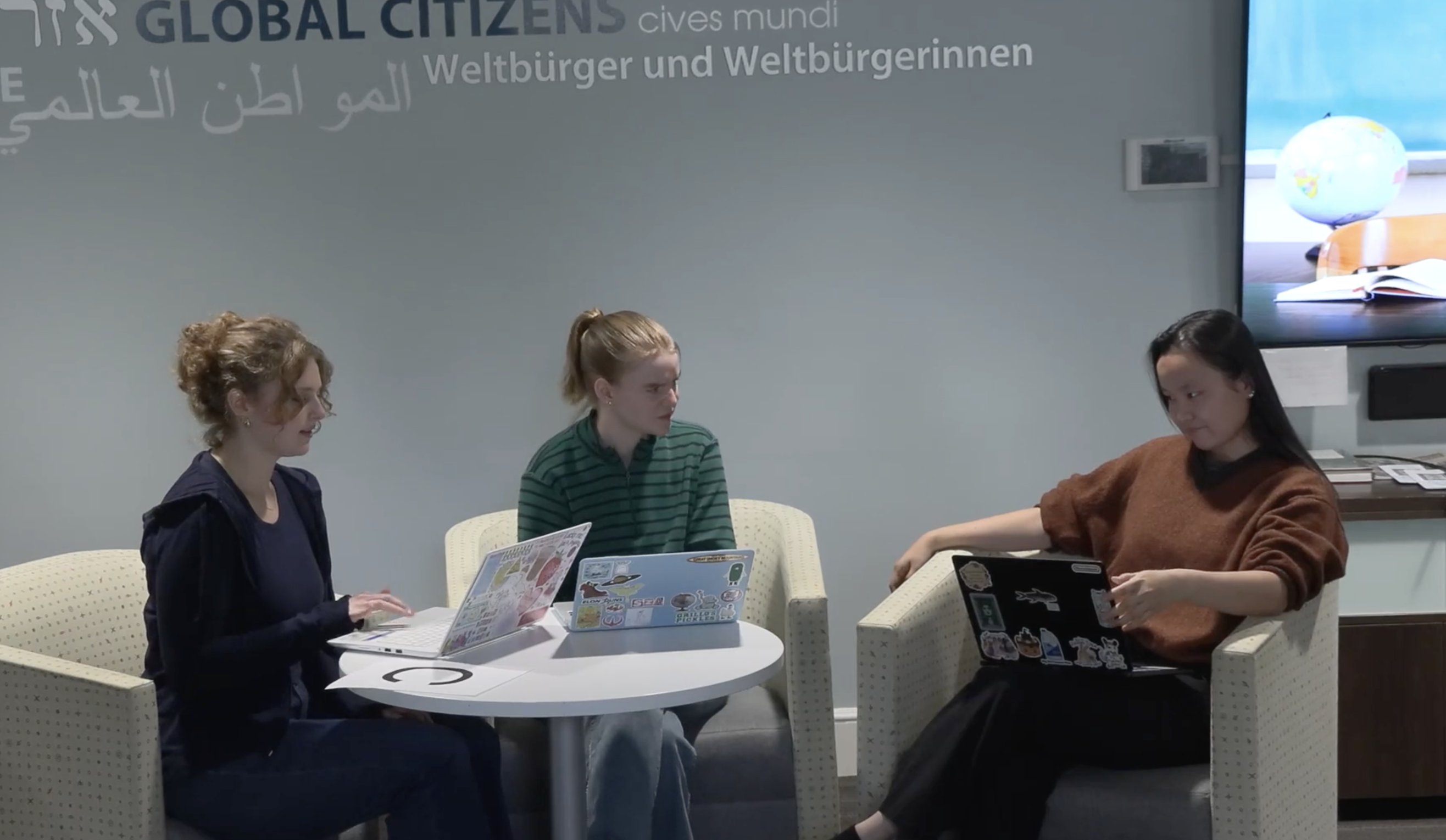 Alt text (two sentences): Three people sit in armchairs around a small table, each working on laptops covered in stickers. Behind them, a wall displays the words “Global Citizens” in multiple languages.